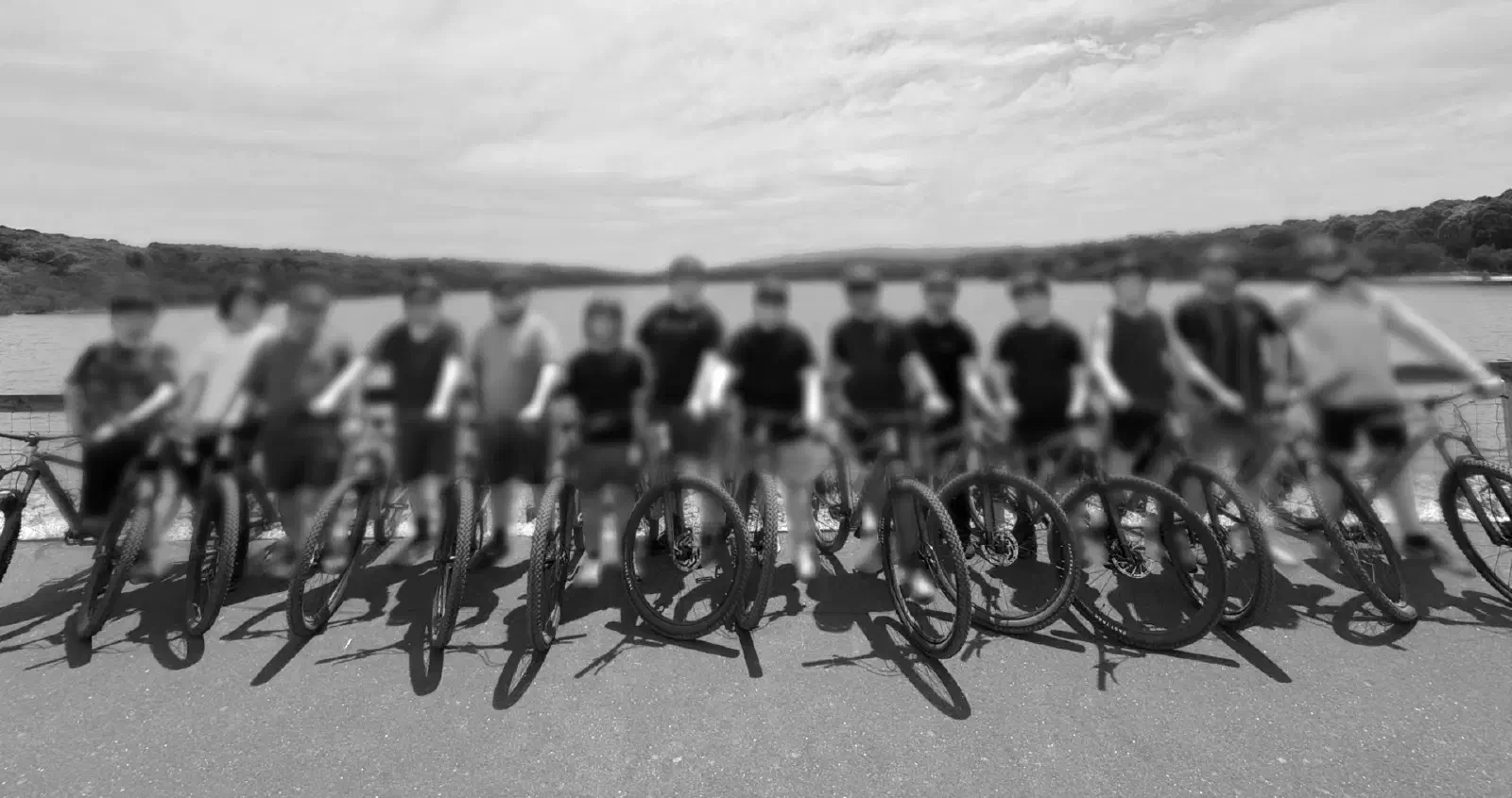 Fourteen people, their faces blurred, standing with mountain bikes in front of a lake