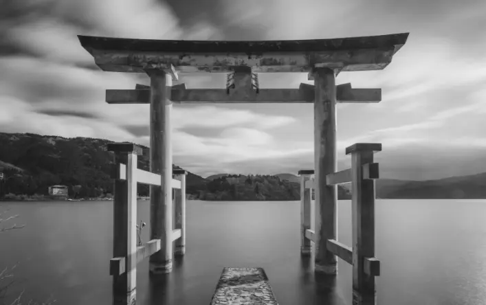 A traditional Japanese gate also known as a torii sitting on the water with mountains and clouds in the background