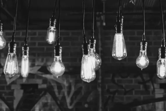 A greyscale shot of old style halogen bulbs hanging from a rustic roof which graffitied bricks in the background.