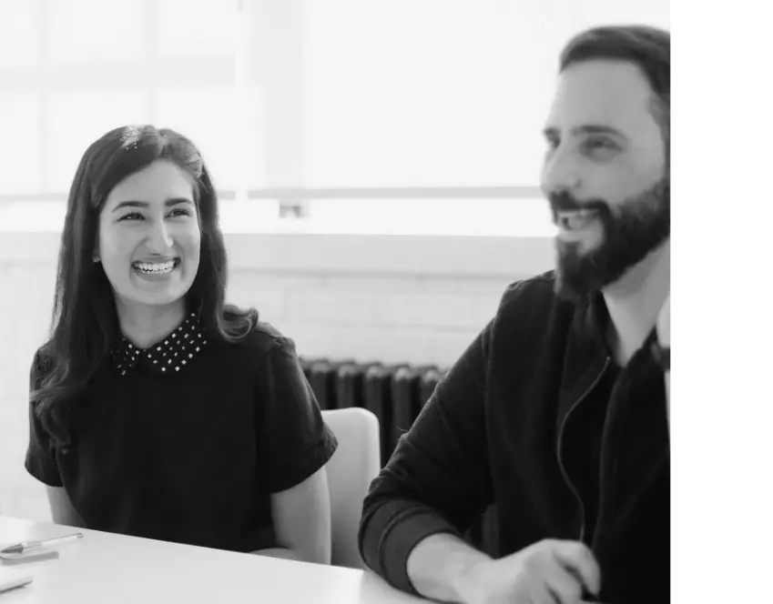 A grayscale shot of a women and man smiling and sitting in smart casual business attire in a meeting.