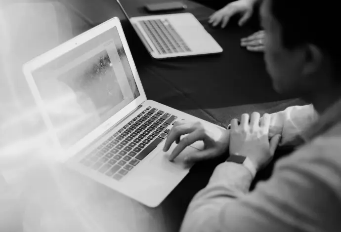 A grayscale shot of a man operating the touchpad of a laptop with a colleague in the background.